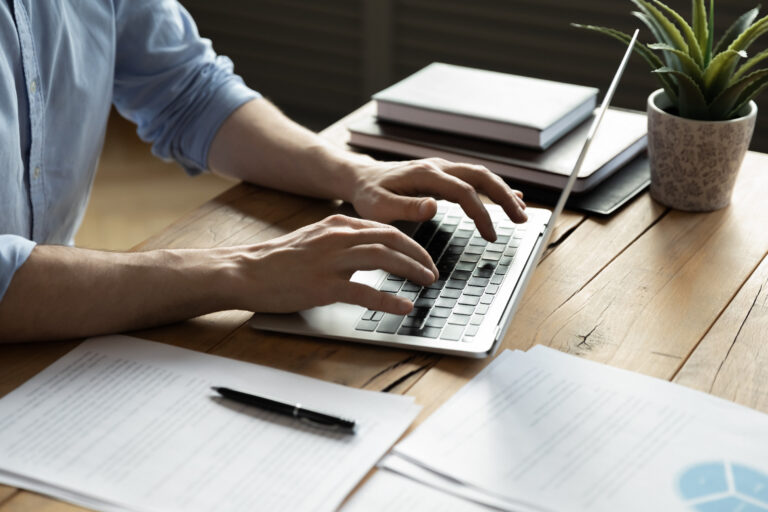 Close up of businessman using laptop, typing on keyboard, sitting at wooden desk with documents